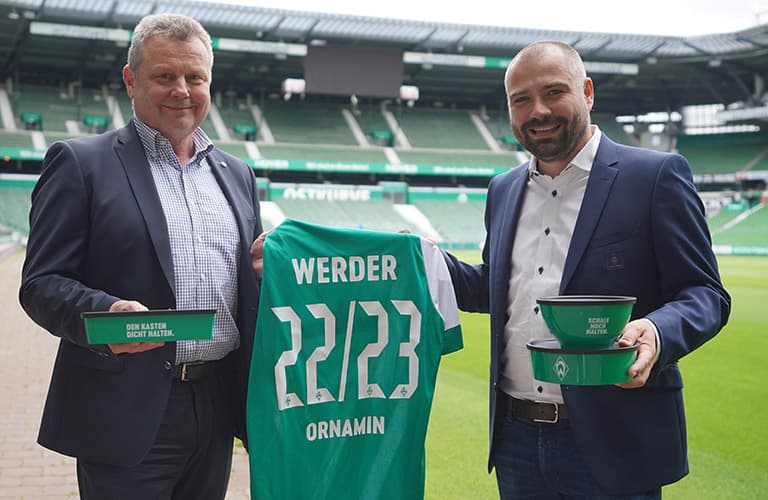 ORNAMIN-Blog-Ornamin ist Supplier des SV Werder Bremen Zwei Männer halten ein Werder Bremen Trikot und Werder Bremen Mehrwegdosen zum Mitnehmen von Essen