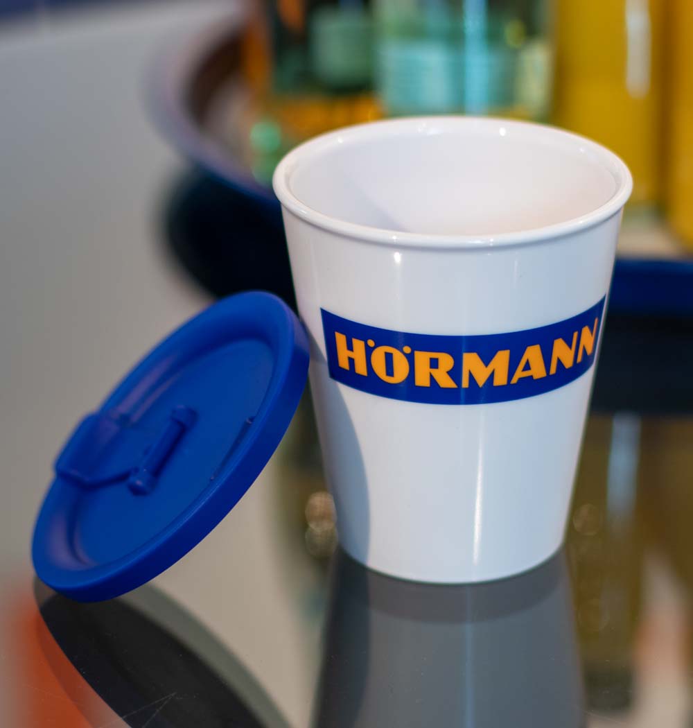 ORNAMIN-Mehrwegsystem-für-Unternehmen-Coffee-to-go-Referenz-Hoermann ORNAMIN Coffee to-go-Mehrwegbecher im Design von Hoermann steht mit angelehntem Deckel auf dem Tisch
