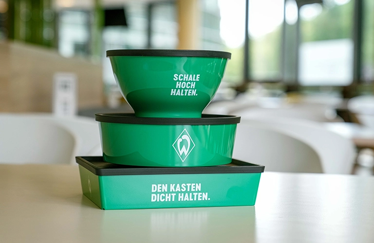 ORNAMIN-Refrenzen-Werder Bremen Mehrwegschalen zum Mitnehmen Auf dem Tisch steht ein Stapel Werder Bremen Mehrwegboxen zum Mitnehmen von Mahlzeiten