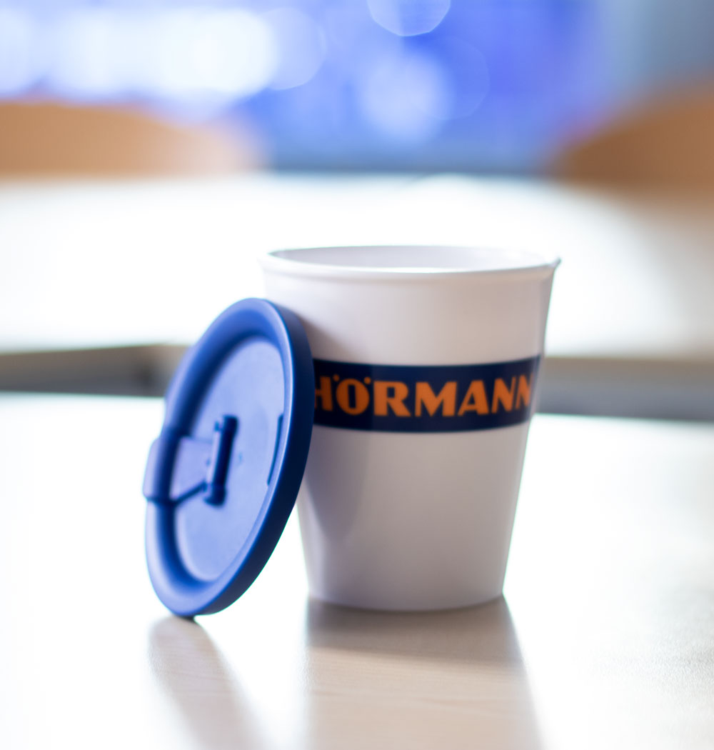 ORNAMIN Coffee to-go Mehrwegbecher im Design von Hoermann steht mit angelehntem Deckel auf dem Tisch