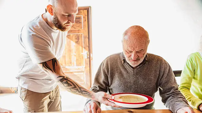 ORNAMIN-Pflegegeschirr-mit-Trick-für-die-professionelle-Pflege Ein Pfleger überreicht pflegebedürftiger Person Suppe im ORNAMIN Kipp-Trick Teller