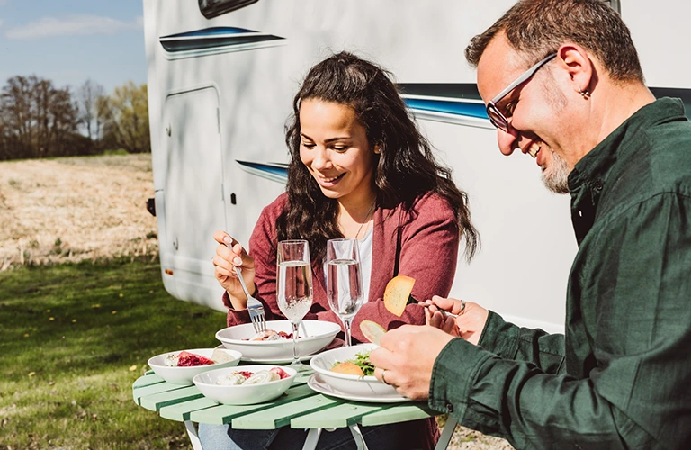 Ein Pärchen beim Essen vor dem Wohnwagen. Auf dem Tisch bruchstabiles Campinggeschirr.