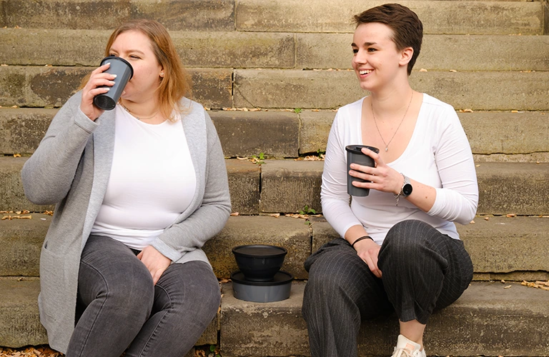 Zwei junge Frauen sitzen im Freien und trinken aus Zero Waste Bechern. 